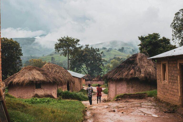 Two men walking through a rural village with thatched roof huts and lush greenery.