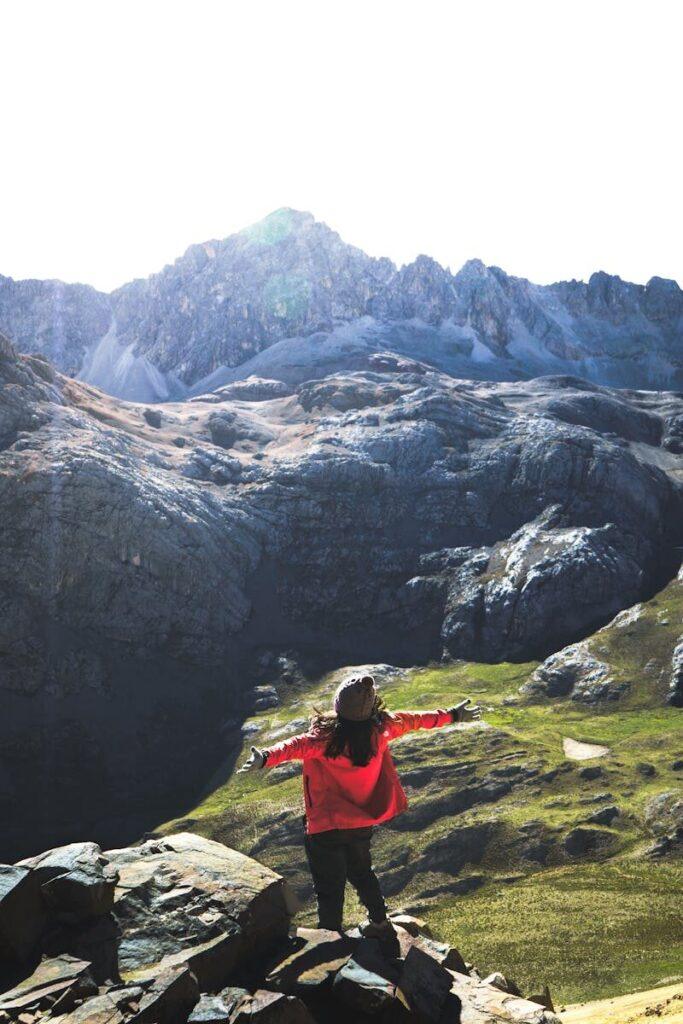 A woman in red stands on rocks, arms open to the majestic Huaral mountains under a bright sky.